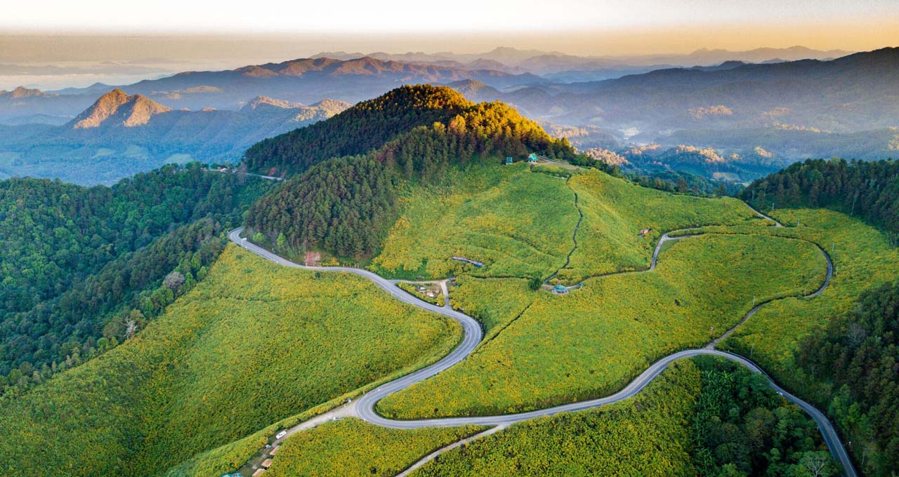 The Mae Hong Son Loop offers one of Southeast Asia’s most scenic road trips, weaving through 1,864 curves, mountain passes, and hill tribe villages in northern Thailand. A winding mountain road surrounded by lush green hills and misty forests along the Mae Hong Son Loop in northern Thailand.
